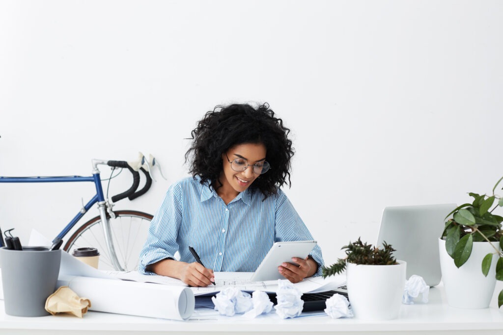 A focused young woman writing at her desk with a tablet, learning how to Become A Junior Bookkeeper Without Matric in a bright and creative workspace.