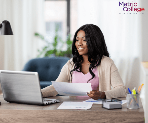 Smiling woman working at her laptop while reviewing documents, representing how students gain Scarce Skills and Their Place In ICB for career success.