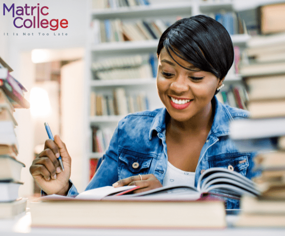 A smiling student studies in a library surrounded by books, representing the opportunity to complete education through Matric College. Where Can I Do Grade 11 And 12?
