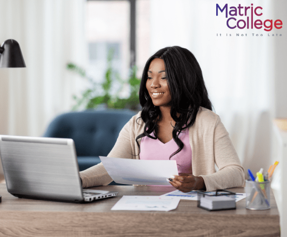Woman sitting at a desk with a laptop and holding documents, smiling while studying, representing the topic "Can I Study ICB Bookkeeping Courses Without Matric?"